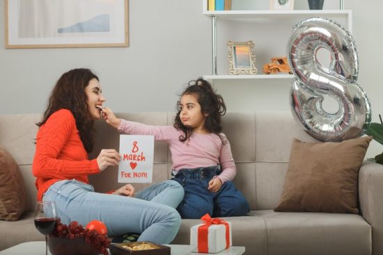 happy-mother-with-her-little-child-daughter-sitting-couch-holding-greeting-card-smiling-cheerfully-light-living-room-celebrating-international-women-s-day-march-8_141793-111611