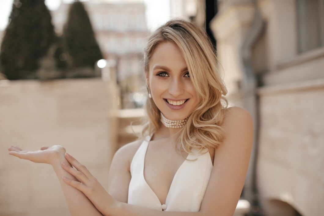 cool-joyful-blonde-woman-v-neck-white-dress-pearl-necklace-smiles-sincerely-looks-into-camera-points-left-street_197531-23188