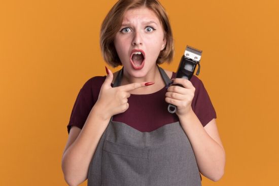 young-beautiful-woman-hairdresser-apron-holding-trimmer-pointing-with-index-finger-it-being-surprised-confused-standing-orange-wall_141793-64846