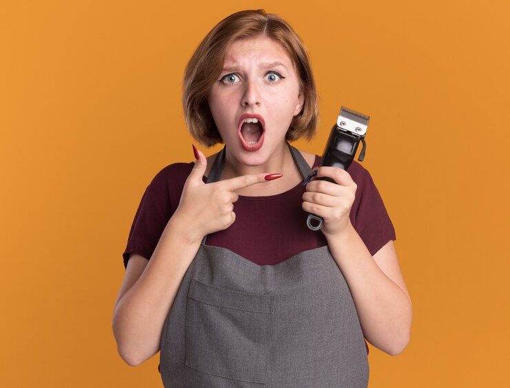 young-beautiful-woman-hairdresser-apron-holding-trimmer-pointing-with-index-finger-it-being-surprised-confused-standing-orange-wall_141793-64846